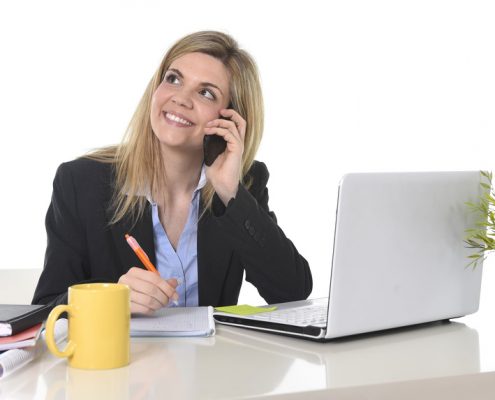 happy Caucasian blond business woman working talking on mobile phone at office computer desk