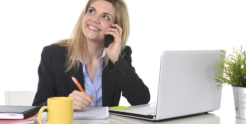 happy Caucasian blond business woman working talking on mobile phone at office computer desk